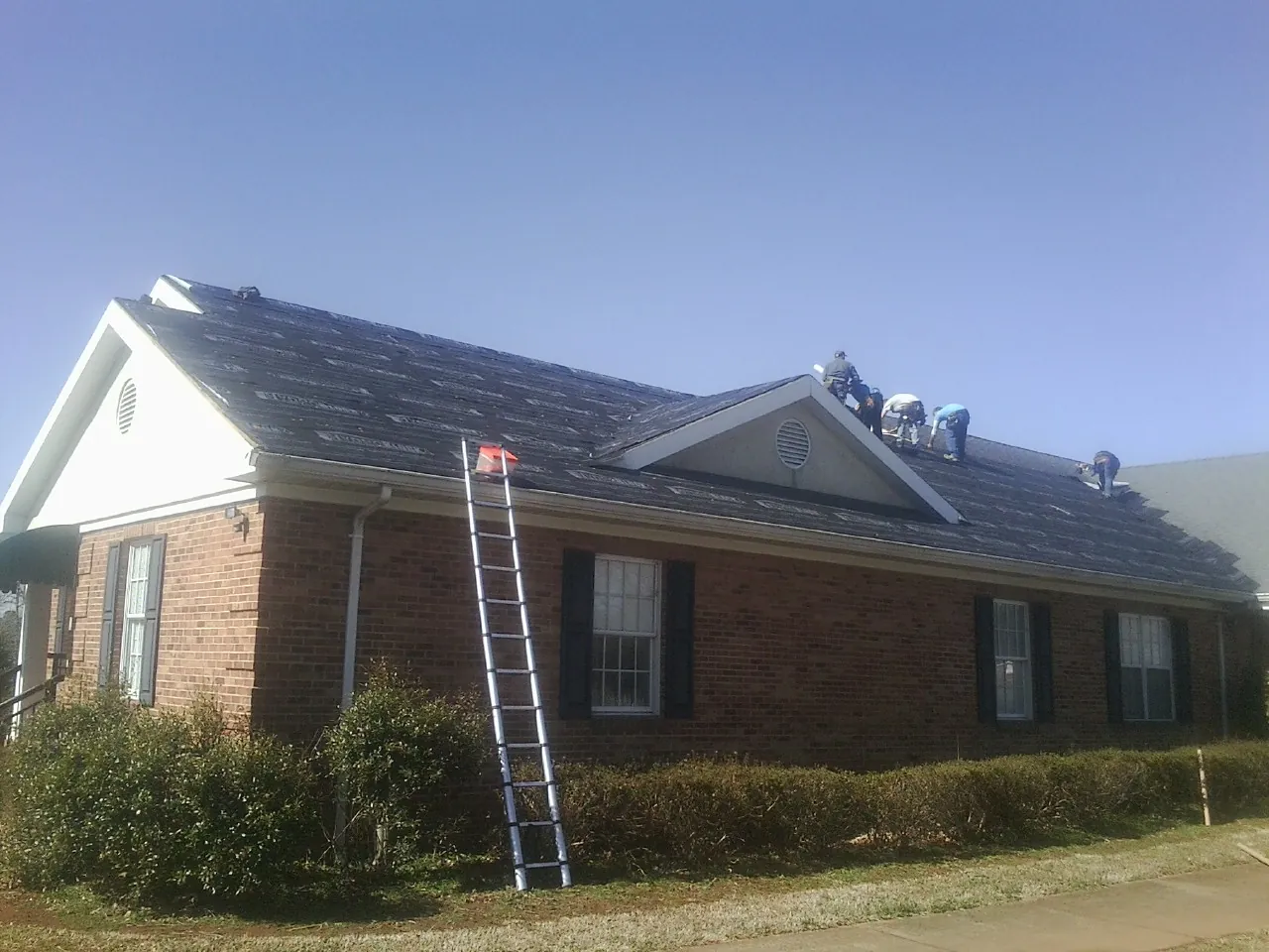 Crew tearing shingles off a brick ranch roof with a ladder set — Metal Roof Repair work by our crew in Fellsmere