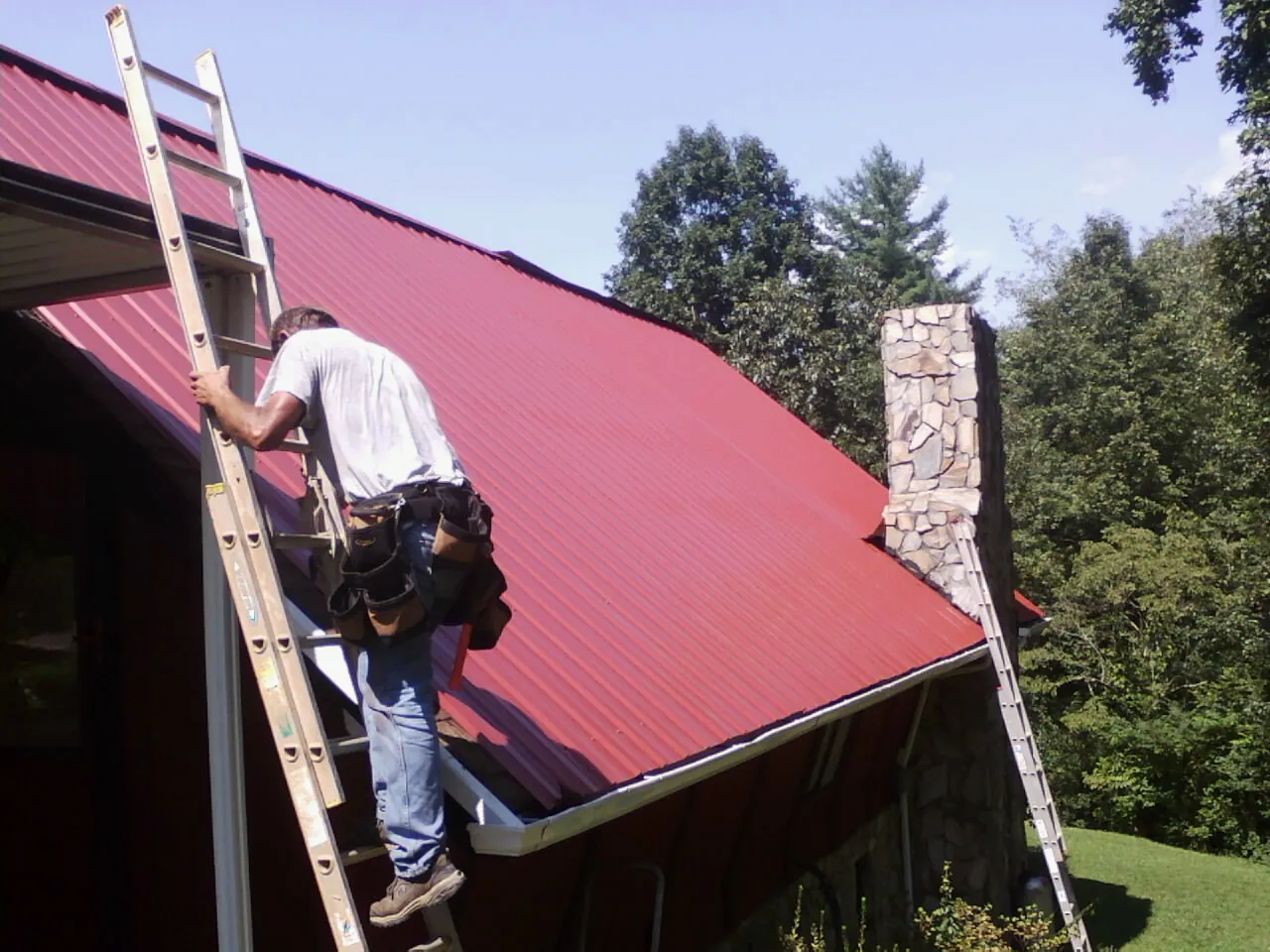 Roofer on a ladder beside a red metal roof and stone chimney — Soffit & Fascia Repair by Roofing Brothers in Fellsmere